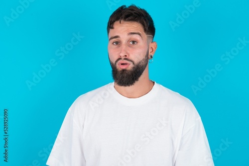 bearded caucasian man wearing white T-shirt over blue background expressing disgust, unwillingness, disregard having tensive look frowning face, looking indignant with something.