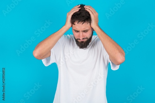 bearded caucasian man wearing white T-shirt over blue background holding head with hands, suffering from severe headache, pressing fingers to temples