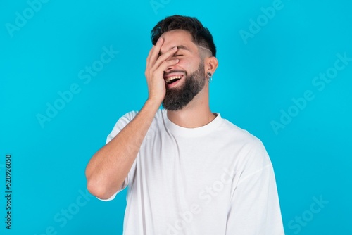 Charismatic carefree joyful bearded caucasian man wearing white T-shirt over blue background likes laugh out loud not hiding emotions giggling hear funny hilarious joke chuckling facepalm.