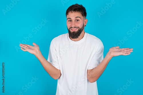 So what? Portrait of arrogant bearded caucasian man wearing white T-shirt over blue background shrugging hands sideways smiling gasping indifferent, telling something obvious.