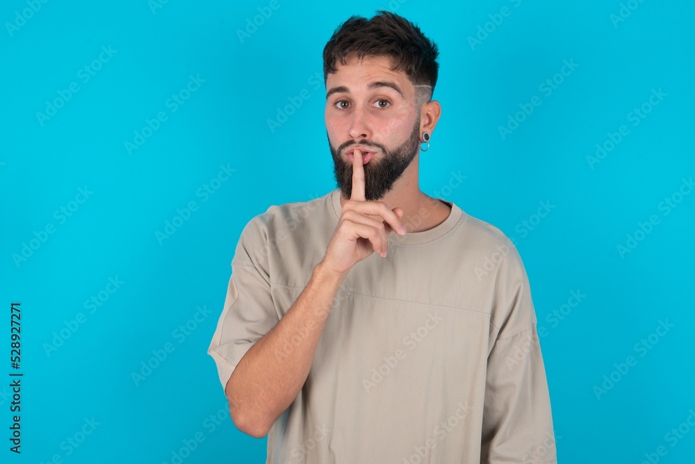 Surprised bearded caucasian man wearing casual T-shirt over blue background makes silence gesture, keeps finger over lips and looks mysterious at camera Surprised bearded caucasian man wearing casual T-shirt over blue background makes silence gesture, keeps finger over lips and looks mysterious at camera