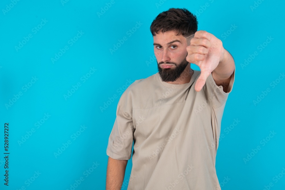 bearded caucasian man wearing casual T-shirt over blue background looking unhappy and angry showing rejection and negative with thumbs down gesture. Bad expression. bearded caucasian man wearing casual T-shirt over blue background looking unhappy and angry showing rejection and negative with thumbs down gesture. Bad expression.