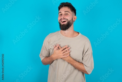bearded caucasian man wearing casual T-shirt over blue background expresses happiness, laughs pleasantly, keeps hands on heart