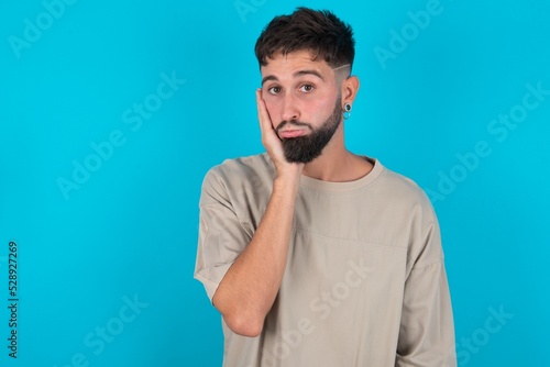 Sad lonely bearded caucasian man wearing casual T-shirt over blue background touches cheek with hand bites lower lip and gazes with displeasure. Bad emotions