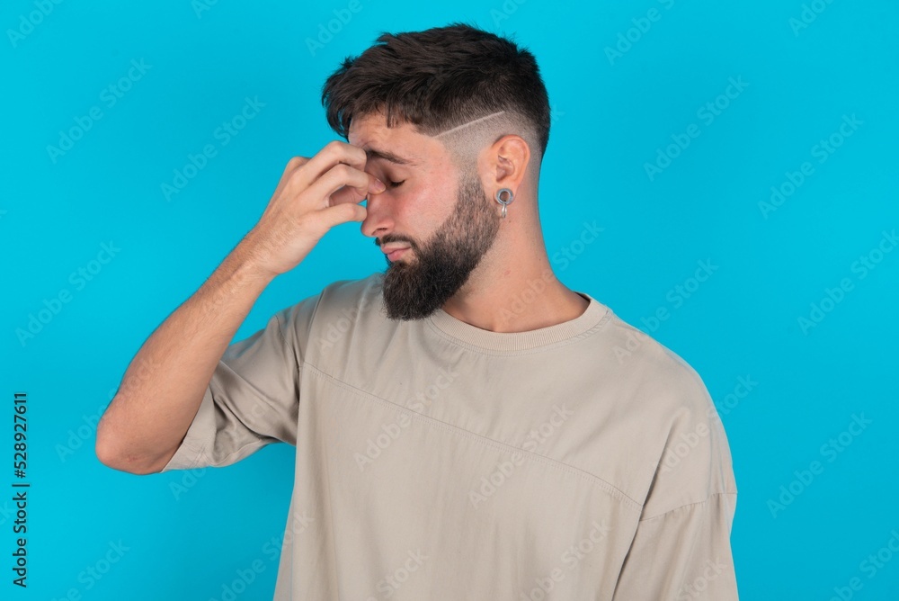 Sad bearded caucasian man wearing casual T-shirt over blue background suffering from headache holding hand on her face Sad bearded caucasian man wearing casual T-shirt over blue background suffering from headache holding hand on her face
