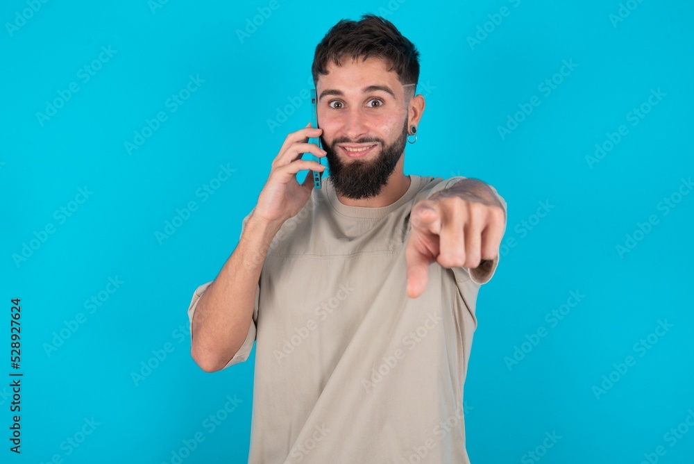 Positive bearded caucasian man wearing casual T-shirt over blue background indicates directly at camera has telephone conversation smiles broadly enjoys talking long hours. You join me Positive bearded caucasian man wearing casual T-shirt over blue background indicates directly at camera has telephone conversation smiles broadly enjoys talking long hours. You join me