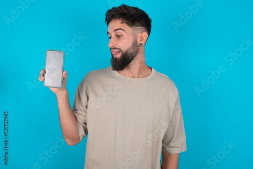Photo of nice pretty bearded caucasian man wearing casual T-shirt over blue background demonstrate phone screen hold hair tails