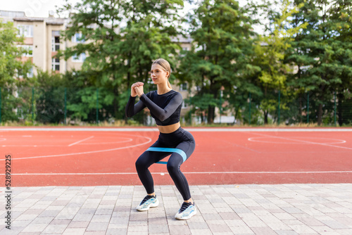 Concept of healthy and active lifestyle. Profile side view of young woman making sport training outdoors alone, squat with resistance band equipment, holding hands in front