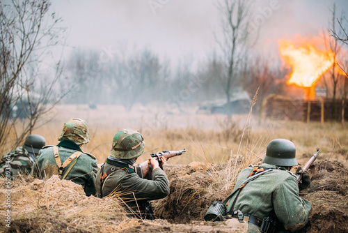 Re-enactors Armed Rifles And Dressed As World War Ii German Wehrmacht Infantry Soldiers Fighting Defensively In Trench. Defensive Position. Smokescreen. Building On Fire On Background.