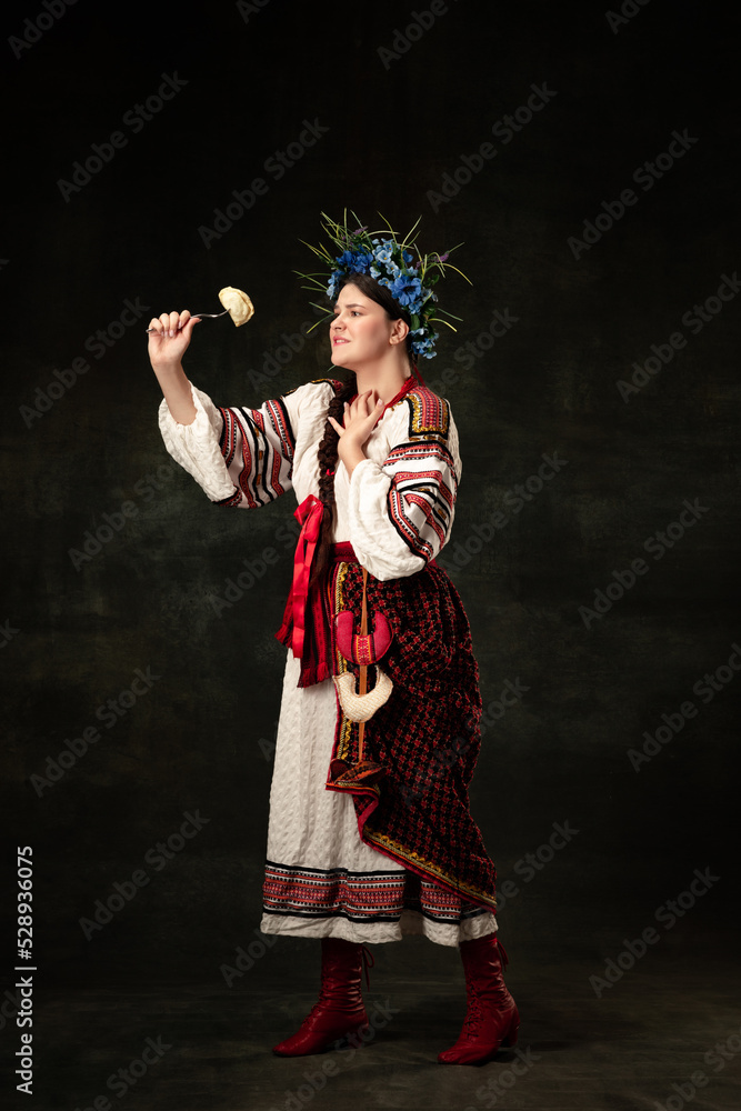 Creative portrait of beautiful Ukrainian woman wearing traditional folk costume tasting varenik isolated over dark background. Traditions, national cuisine