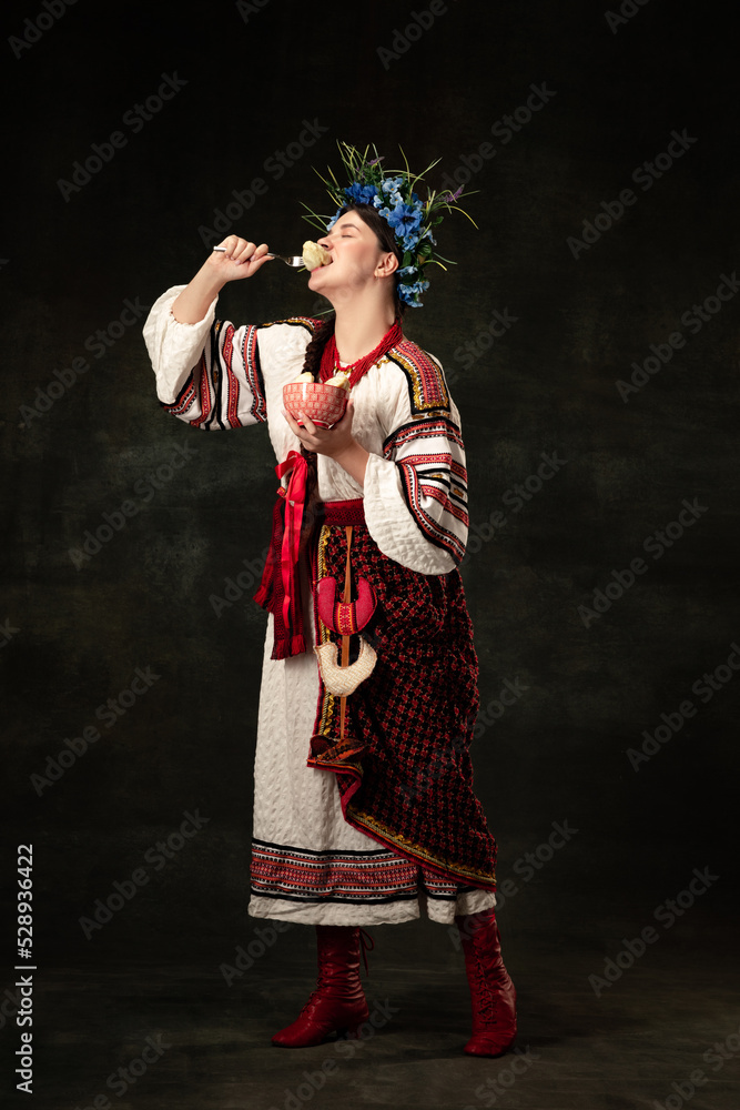Enjoy. Creative portrait of beautiful Ukrainian woman wearing traditional folk costume tasting varenik isolated over dark background.