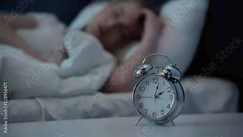 Alarm clock on bedside table near blurred woman lying in bedroom at night.