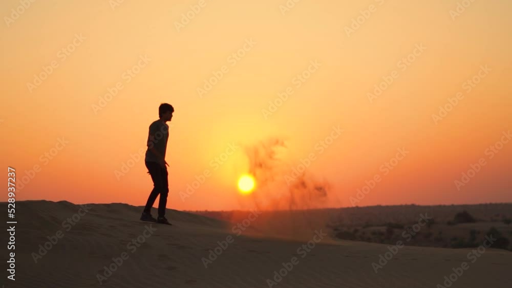Slow motion silhouette shot of an Indian man kicking the sand of the Sam sand dunes of Thar desert in front of the sun during the sunrise at Jaisalmer in Rajasthan, India. Tourist playing with sand. Slow motion silhouette shot of an Indian man kicking the sand of the Sam sand dunes of Thar desert in front of the sun during the sunrise at Jaisalmer in Rajasthan, India. Tourist playing with sand.