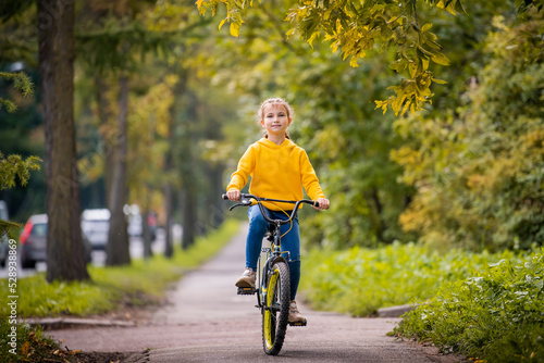 smiling girl of ten in a yellow sweater rides bicycle on autumn street under the branches of a red rowan tree. © Maria Moroz