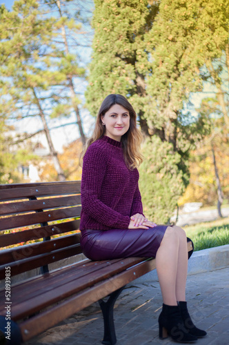 Beautiful woman in burgundy sweater sitting on the bench on a background of autumn tree.
