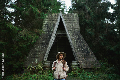 Millennial traveler woman in hat with backpack standing in front of old vintage style wooden hut in the dark forest photo