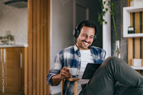 A happy man spending time at home, working over the tablet, drinking coffee, and listening to music over headphones.