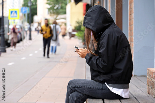 Girl wearing black coat with hood and jeans sitting with smartphone and cigarette on а street on people background. Smoking and using mobile phone in autumn city