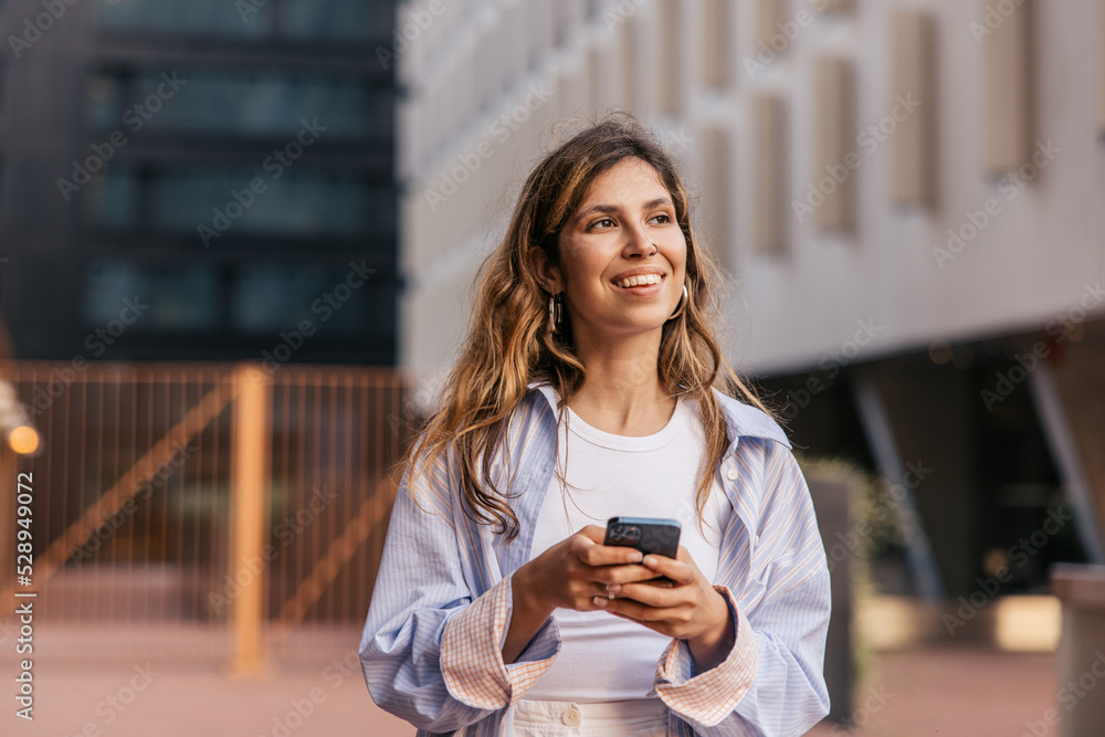 Fototapeta premium Beautiful young caucasian girl uses smartphone spending leisure time on street. Blonde smiles looking away, wears casual clothes. City life concept