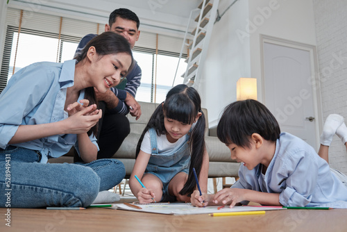 Asian Thai siblings and mum are sitting on living room floor, drawing with colored pencils together, dad leisurely relaxing on a sofa, lovely happy weekend activity, and domestic wellbeing lifestyle. © tigercat_lpg