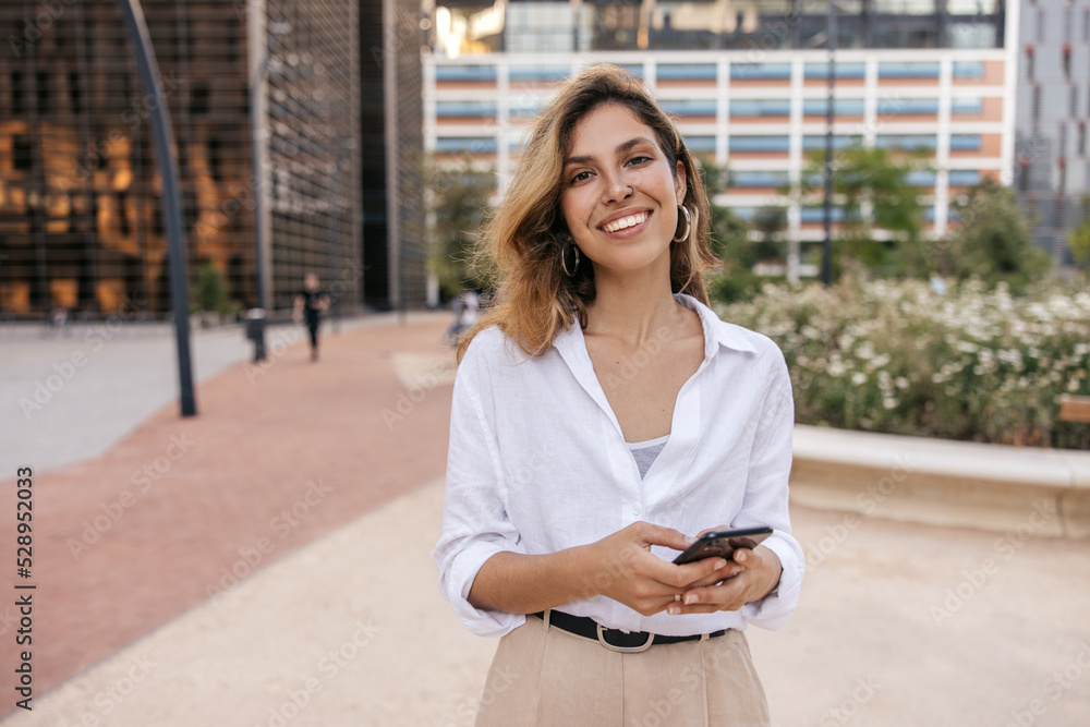 Fototapeta premium Smiling young caucasian woman is using smartphone, city with skyscrapers in background. Blonde smiles, wears classic style of clothing. Technology concept