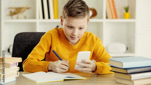 schoolboy looking at smartphone while writing in notebook near books in classroom. photo