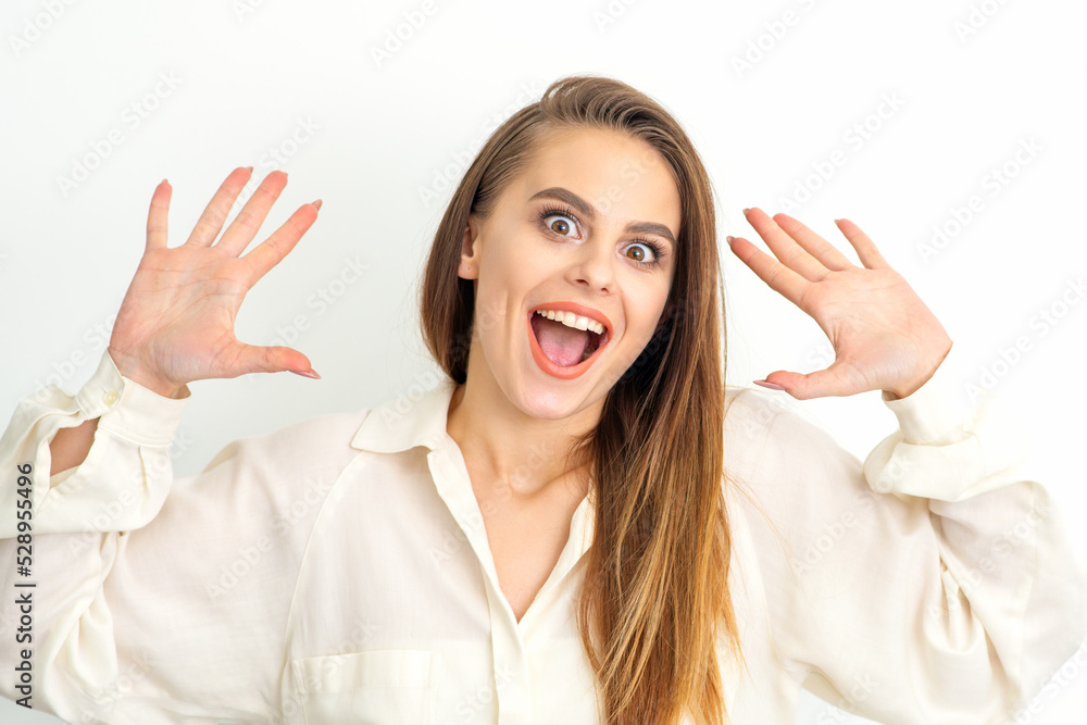 Portrait of young caucasian woman wearing white shirt raises hands and laughs positively with open mouth poses against a white background Portrait of young caucasian woman wearing white shirt raises hands and laughs positively with open mouth poses against a white background