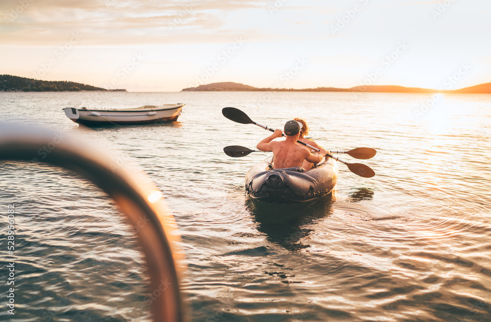 Two rowers on inflatable kayak rowing by the evening sunset rays Adriatic sea harbor in Croatia near Sibenik city. Vacation, sports, and a recreation concept image. - obrazy, fototapety, plakaty 