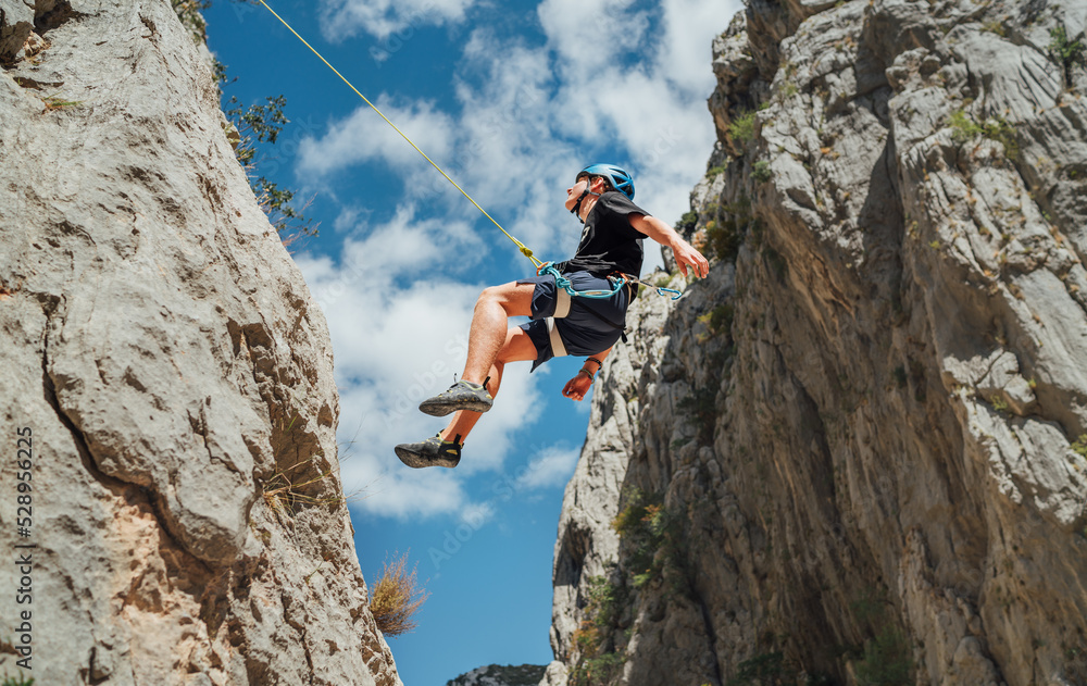 Obraz premium Climber teenager boy in protective helmet abseiling from vertical cliff rock wall using rope Belay device, climbing harness in Paklenica canyon in Croatia. Active extreme sports time spending concept