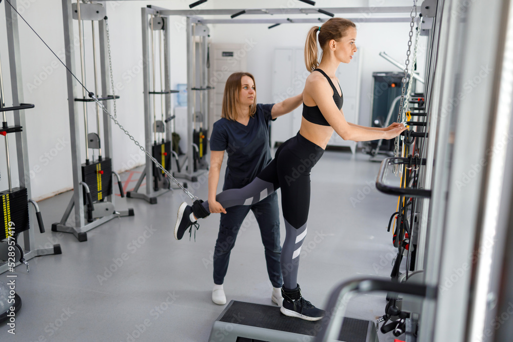 Fototapeta premium Young woman doing rehabilitation exercises with personal instructor on the kinesi machine