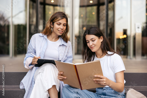 Two caucasian young female friends study using books together sitting on stairs outdoors. Blonde and brunette wear casual clothes. University life on campus.