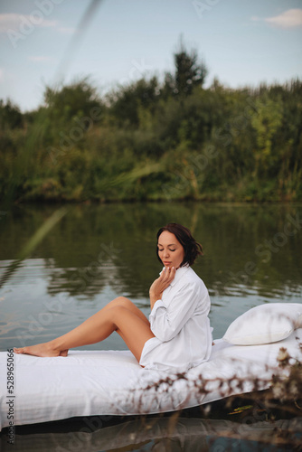 Brunette woman with short hair in a white shirt in bed on the water. A young woman floats on an inflatable mattress with a white sheet on the lake. © VikaNorm