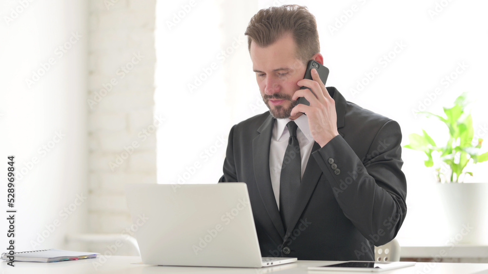 Young Businessman Talking on Phone while using Laptop in Office Young Businessman Talking on Phone while using Laptop in Office