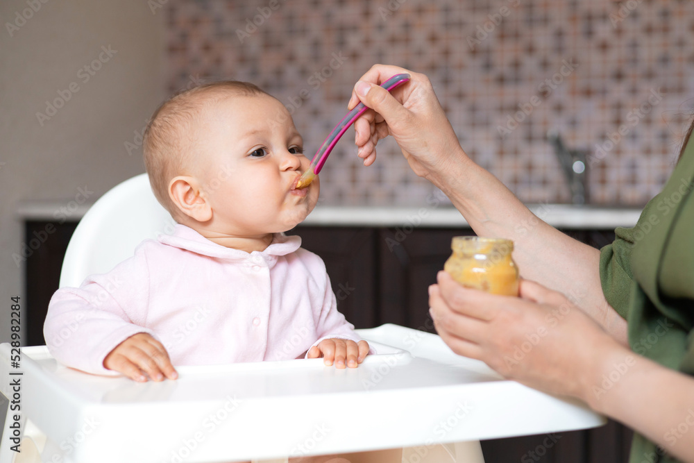 Baby food. A baby in a high chair eats vegetable puree from a spoon. Mom feeds the baby from a jar. Kitchen. Lifestyle. Baby food. A baby in a high chair eats vegetable puree from a spoon. Mom feeds the baby from a jar. Kitchen. Lifestyle.