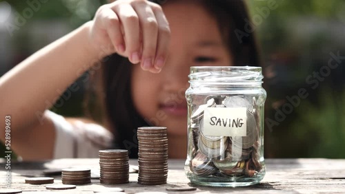 Happy Asian child girl save money by putting coins in glass jar, coin stack growing. Concept saving money, cash, education, growth, finance, bank, investment, management, income, business