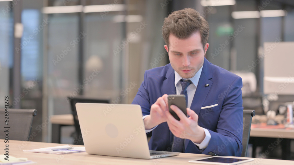 Businessman using Smartphone while using Laptop in Office Businessman using Smartphone while using Laptop in Office