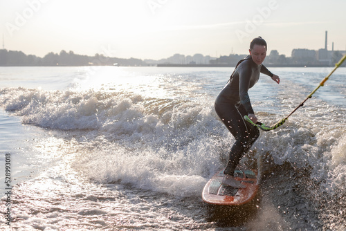 An active athletic woman in a wetsuit rides a splashing river wave on a wakesurfing board with cable in her hands photo