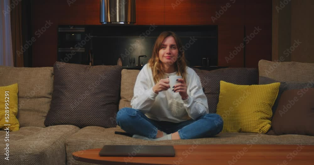 Young woman turns on TV, watching television, drinking tea, smiling, sitting on sofa on evening at home Young woman turns on TV, watching television, drinking tea, smiling, sitting on sofa on evening at home