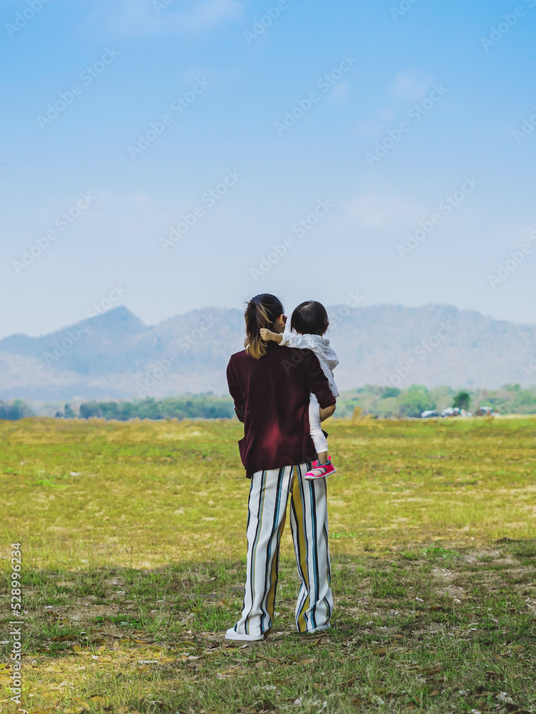 Back view of happiness Asian mother holding daughter with meadow and mountain in background. Lovely Family looking at beautiful sky together. Joy of outdoor living together. Happy family relationship.