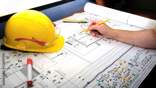 An engineer is working on blueprints. A yellow hard hat and goggles lie on an engineering diagram of an energy industrial facility with pencils and a marker.