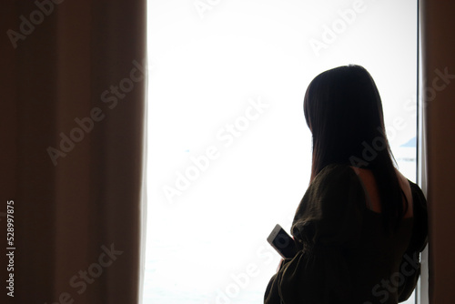 Back side of Woman holding cellphone standing near window looking outside. Woman pose indoor distracted from who is quarantined at home due to covid-19. photo