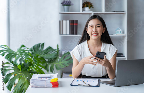 Beautiful Asian businesswoman using smartphone at office desk, female accountant working on laptop, financial audit concept.