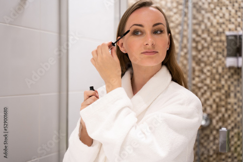 Young woman in white robe paints lashes with mascara looking in mirror. Lady stands in hotel bathroom doing daily makeup and smiling close view