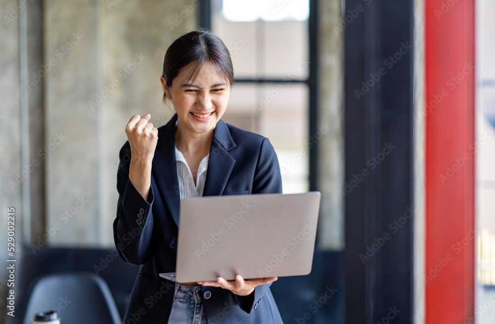 Happy excited successful Asian businesswoman triumphing with a laptop computer smartphone in the workplace office Happy excited successful Asian businesswoman triumphing with a laptop computer smartphone in the workplace office