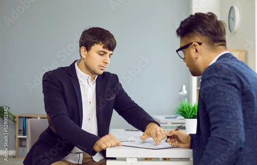 Quadro em tela Two serious business people in office sign a contract sitting at desk, manager gives document to his client