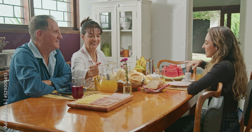Happy couple having breakfast with teenage daughter sitting at kitchen table. The father is talking and the mother is serving juice to the daughter Happy couple having breakfast with teenage daughter sitting at kitchen table. The father is talking and the mother is serving juice to the daughter