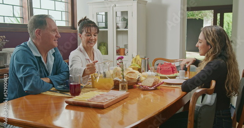 Happy couple having breakfast with teenage daughter sitting at kitchen table. The father is talking and the mother is serving juice to the daughter