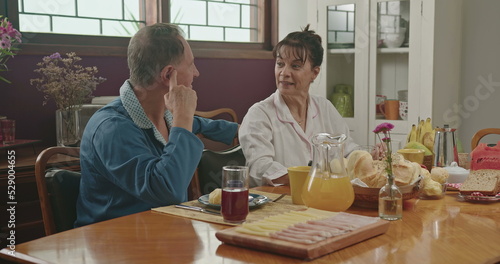 A middle aged couple chatting during breakfast. They are sitting at the lavish kitchen table, smiling and wearing pajamas. Married couple in conversing in the morning breakfast table