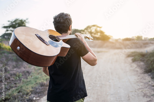 Young musician walking on a countryside road with a guitar on his shoulder