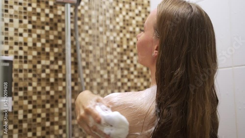 Young woman rubs body with loofah spreading gel foam on shoulders. Lady enjoys taking morning shower after waking up in hotel bathroom closeup photo